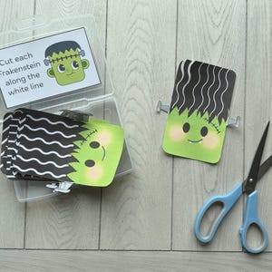 May include: A clear plastic container holds a stack of Frankenstein-themed cards with black wavy hair and green faces. One card is separate, and a pair of blue scissors rests nearby. The text on the card reads: "Cut each Frankenstein along the white line."