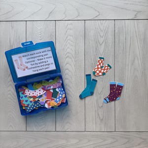 Op de afbeelding: Een blauwe plastic doos met kleurrijke sokuitsparingen, met drie paar sokken die buiten worden getoond. Het deksel van de doos heeft de tekst: "Combineer elke sok met het bijbehorende paar." De sokken hebben verschillende patronen en kleuren.