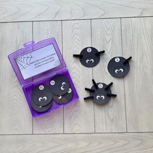 May include: A purple container with a matching lid holds black spider-shaped game pieces with numbers 1, 2, 5, 6, and 8. The game pieces have googly eyes and black pipe cleaner legs. The text on the lid reads, "Match the number of clothes pins to corresponding number on each spider."