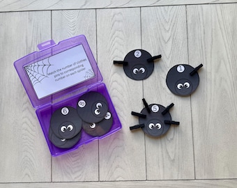 May include: A purple container with a lid holds black spider-shaped game pieces with numbers 1, 2, 5, 6, and 8. The game pieces have googly eyes and black pipe cleaner legs. The text on the lid reads, "Match the number of clothes pins to corresponding number on each spider."