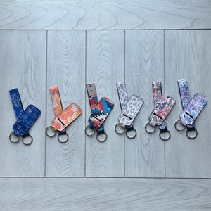 May include: A collection of keychains with various patterns, including floral, geometric, and animal print designs. Each keychain features a matching wristlet strap and a metal keyring. The color palette includes blue, orange, red, white, and pink.