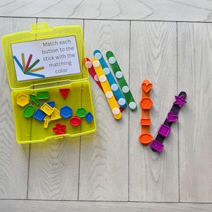 May include: A yellow plastic container with colorful buttons and matching sticks. The container has a label that reads, "Match each button to the stick with the matching color." The buttons are various shapes and colors, and the sticks are decorated with white dots.