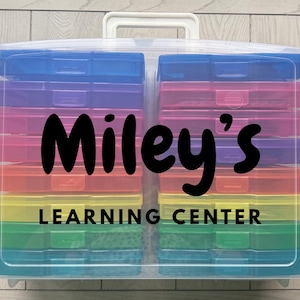 May include: A clear plastic storage container with a white handle, filled with rainbow-colored compartments. The text "Miley's LEARNING CENTER" is displayed in black on the front of the container.