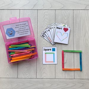 Puede incluir: Un recipiente de plástico rosa lleno de palitos de helado de colores, tarjetas de tareas y un cuadrado construido. Las tarjetas de tareas muestran formas como un corazón y un cuadrado. El texto en el contenedor dice: "Construye la forma que se muestra en cada tarjeta de tareas con palitos de helado con el palito de color correspondiente."