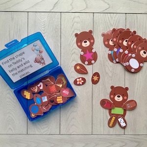 Può includere: Una scatola di plastica blu con pezzi di puzzle a forma di orso marrone. Il testo sul coperchio recita: "Trova la forma sulla pancia di Teddy e aggiungi i pezzi corrispondenti". Diversi ritagli di orsi e forme corrispondenti sono sparsi.