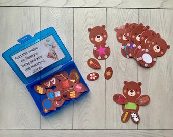 May include: A blue plastic container with a lid, filled with brown bear-shaped puzzle pieces. The text on the lid reads "Find the shape on Teddy's belly and add the matching pieces." Several bear cutouts and matching shapes are scattered around the container.