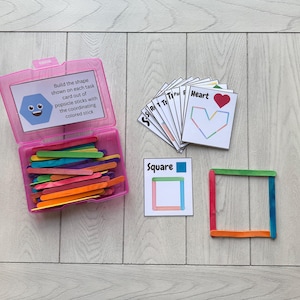 Peut inclure: Une boîte en plastique rose contenant des bâtonnets de glace colorés, des cartes de tâches et un carré construit. Les cartes de tâches indiquent aux utilisateurs de construire des formes avec les bâtonnets. Le texte sur le conteneur se lit comme suit : "Construisez la forme indiquée sur chaque carte de tâche avec des bâtonnets de glace avec le bâtonnet coloré correspondant."