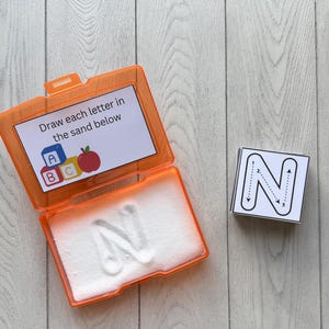 May include: An orange plastic case with a white sand tray and the letter "N" imprinted in the sand. A card with the text "Draw each letter in the sand below" is visible. A separate card shows the letter "N" with directional arrows.
