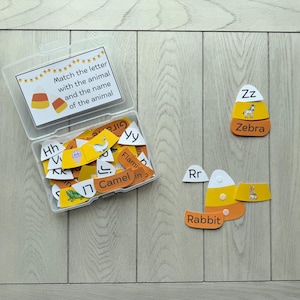 Puede incluir: Una caja de almacenamiento de plástico transparente con tarjetas educativas en forma de maíz dulce que muestran letras, ilustraciones de animales y nombres. La tapa tiene el texto Match the letter with the animal and the name of the animal. Dos tarjetas fuera.