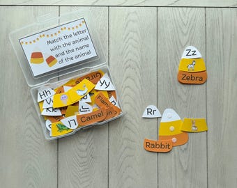 May include: A clear plastic storage box containing candy corn-shaped educational cards with letters, animal illustrations, and names. The box lid features the text Match the letter with the animal and the name of the animal. Two assembled cards are outside.