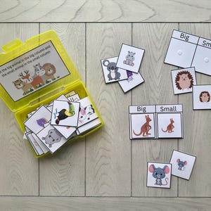 Może przedstawiać: Żółte plastikowe pudełko z pokrywką, wypełnione kartami ze zwierzętami do gry w sortowanie. Karty zawierają ilustracje różnych zwierząt, a gra zawiera karty z etykietami "Duży" i "Mały".