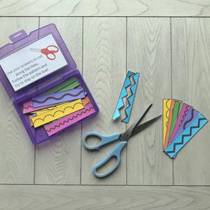 以下が含まれることがあります： カラフルな紙のストリップとハサミが入った紫色のプラスチック容器。容器には、「ハサミを使って線に沿ってカットしてください。パターンに従い、線上に留まるようにしてください！」と書かれた白いラベルが付いています。ハサミは青色です。