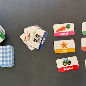 Puede incluir: Juego de tarjetas ilustradas con palabras e imágenes, como zanahoria, estrella, tractor y oveja. Dispuestas sobre una superficie oscura, con una pila a cuadros azules y blancos y una caja azul.