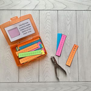 Pode incluir: Uma caixa de plástico laranja aberta contendo tiras de papel coloridas com círculos pré-impressos. Um furador e tiras adicionais estão ao lado da caixa. O texto na caixa diz: "Use o furador para fazer furos em todos os pontos da tira de papel."