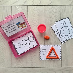 以下が含まれることがあります： ピンク色のプラスチック容器に入ったタスクカードと粘土。カードには粘土で埋める形があります。カードのテキストは「私は...を作ることができます」と「各タスクカードに表示されている形に粘土を転がします」と書かれています。
