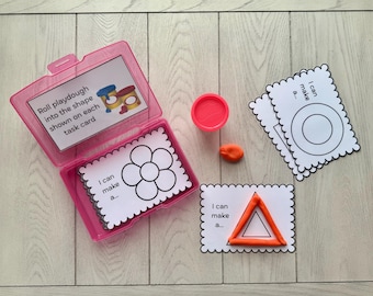 May include: A pink plastic container with task cards and playdough. The cards have shapes to be filled with playdough. The text on the cards reads "I can make a..." and "Roll playdough into the shape shown on each task card."