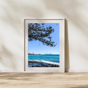 Puede incluir: Fotografía enmarcada de una escena costera. La imagen presenta un océano azul vibrante, una costa rocosa y un cielo azul claro. Una rama de árbol se extiende hacia el marco desde la parte superior izquierda. Se ven edificios en el horizonte.