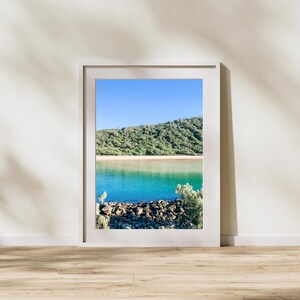 Könnte beinhalten: Gerahmte Landschaftsfotografie mit einer Küstenlandschaft. Das Bild zeigt einen klaren blauen Himmel, türkisfarbenes Wasser, einen Sandstrand und einen grünen Hügel. Im Vordergrund befindet sich eine Felsbarriere.