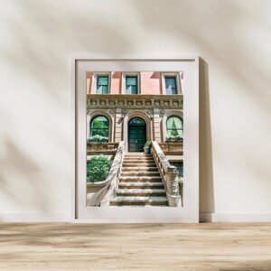 Puede incluir: Una impresión enmarcada de un edificio de piedra rojiza con una puerta verde oscuro y escaleras que conducen a ella. El edificio tiene ventanas arqueadas y detalles decorativos. La imagen está colocada contra una pared blanca y un suelo de madera.