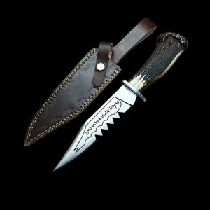 May include: A hunting knife with a serrated blade and a deer antler handle, resting next to a brown leather sheath. The blade has etched text. The sheath has decorative stitching and a leather strap with a button closure. The knife is set against a black background.