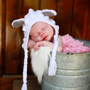 May include: A baby wearing a white crocheted lamb hat with gray ears and a pink ruffled dress is sleeping in a metal bucket. The bucket is sitting on a wooden surface.