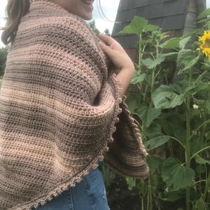 May include: A crocheted shawl with a brown and pink ombre color pattern. The shawl has a scalloped edge and is worn by a person standing in a garden.