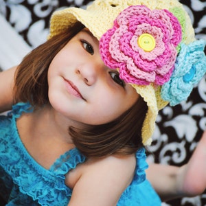 May include: A young girl wearing a yellow crocheted sun hat with pink and blue crocheted flowers. The hat has a wide brim and a button on the front. The girl is wearing a blue ruffled dress.