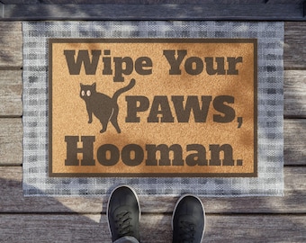 May include: A rectangular doormat with the text "Wipe Your PAWS, Hooman." in brown lettering. A black cat silhouette is between "Your" and "PAWS." The mat has a tan center and a grey plaid border. Black shoes are visible.