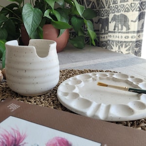 May include: A white ceramic water cup with a notched rim and a matching circular paint palette with multiple indentations, both featuring a speckled finish. A thin paintbrush with a gold ferrule rests across the surface of the paint palette.