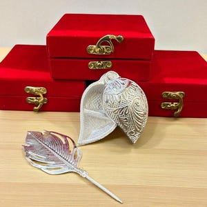 May include: A silver leaf-shaped trinket box with intricate embossed detailing, open to reveal a small compartment. A matching silver feather-shaped pen lies nearby. Three red velvet boxes with gold clasps are in the background.