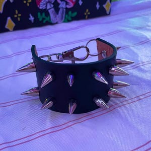 May include: Black leather bracelet with silver cone studs. The bracelet features a metal clasp and a brown leather interior. The bracelet is on a white and pink striped surface.