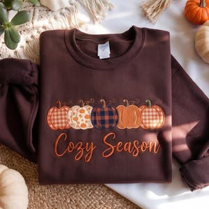 Pode incluir: Uma camisola de gola redonda castanho-escura com uma fila bordada de cinco abóboras com vários padrões, incluindo xadrez e laranja sólido, acima do texto "Cozy Season" numa fonte laranja cursiva. A peça de vestuário tem mangas compridas.