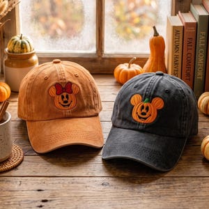 Peut inclure: Deux casquettes de baseball, une beige et une gris anthracite, arborant chacune une citrouille brodée en forme de tête de souris. La casquette beige présente une citrouille avec un nœud rouge, celle en gris montre une citrouille à tige verte.