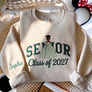 Op de afbeelding: Een beige sweater met ronde hals en donkergroene geborduurde letters met de tekst SENIOR en Class of 2027. In het midden staat een geborduurde afbeelding van een persoon met een afstudeerhoed en een lichtgroene jurk. De naam Sophia staat op de mouw.