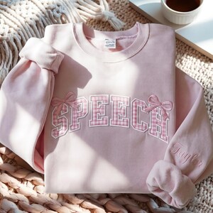 Puede incluir: Una sudadera de cuello redondo rosa claro con la palabra SPEECH en letras de bloque de cuadros vichy rosas con borde blanco en el pecho. El diseño incluye dos lazos rosas bordados en los extremos del texto y el nombre Taylor en la manga.