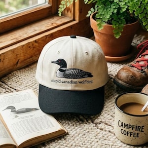 May include: A cream-colored baseball cap with a black brim featuring a graphic of a loon and the text "stupid canadian wolf bird." The cap is next to a mug of coffee and an open book.