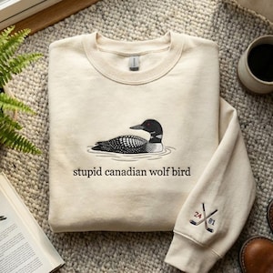 May include: Cream-colored sweatshirt featuring a black and white loon graphic with the text "stupid canadian wolf bird." The sleeve displays crossed hockey sticks with the numbers "24" and "81." The sweatshirt is displayed on a textured surface.