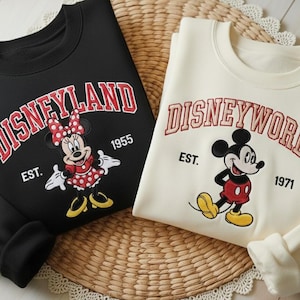 May include: Two sweatshirts, one black and one cream-colored, featuring Disney characters. The black sweatshirt displays "Disneyland" with Minnie Mouse, and the cream one says "Disneyworld" with Mickey Mouse. Both have the establishment year.