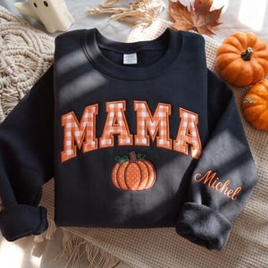 Pode incluir: Uma camisola preta de gola redonda com a palavra MAMA em letras maiúsculas axadrezadas a laranja e branco no peito. Abaixo do texto encontra-se um gráfico bordado de uma abóbora laranja. O nome Michel está bordado em letra cursiva laranja na manga.