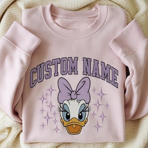 Peut inclure: Un sweat-shirt rose clair avec un motif de Daisy Duck, un nœud violet et le texte "CUSTOM NAME" en violet. Le motif comprend des étoiles. Le sweat-shirt est en matière douce.
