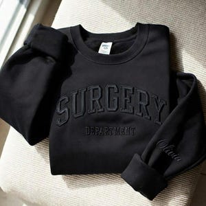 Puede incluir: Una sudadera negra con la palabra "SURGERY" arqueada en el pecho, con "DEPARTMENT" debajo. La manga tiene bordado el nombre "Olivia". La sudadera está doblada sobre una superficie de color claro.