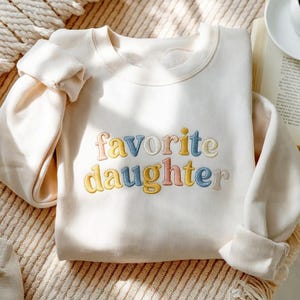 May include: A cream-coloured sweatshirt featuring the embroidered phrase "favorite daughter" in pastel shades. The long-sleeved top is neatly folded. A coffee cup and book are visible in the background.