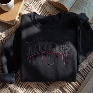 May include: A dark gray crewneck sweatshirt featuring the word RADIOLOGY embroidered in a collegiate font. The text has a red dripping effect underneath, surrounded by embroidered spiderwebs and small flying bats across the chest area.