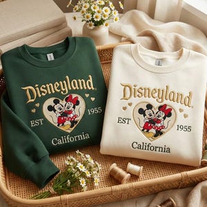 May include: Two sweatshirts, one dark green and one cream, with the Disneyland logo embroidered in gold. The logo includes Mickey and Minnie Mouse in a heart, with "EST 1955 California" below. The sweatshirts are displayed in a woven tray.