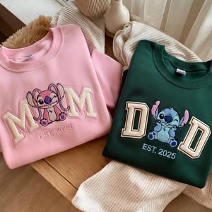 Puede incluir: Dos sudaderas, una rosa y otra verde oscuro, con dise&ntilde;os bordados. La sudadera rosa presenta la palabra "MOM" con un personaje de dibujos animados y "EST. 2025". La sudadera verde dice "DAD" con un personaje de dibujos animados y "EST. 2025".