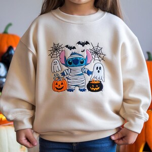 May include: Cream-colored sweatshirt featuring a Halloween-themed graphic. The design includes a blue alien wrapped in bandages, two ghosts, bats, and jack-o'-lanterns. The sweatshirt is a long-sleeved, crew neck style.