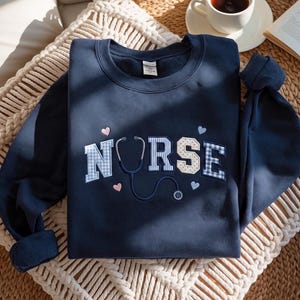Puede incluir: Una sudadera azul marino de cuello redondo con la palabra NURSE bordada en el pecho. Las letras tienen patrones de cuadros vichy azules y lunares beige. Un estetoscopio azul oscuro reemplaza la letra U, decorado con pequeños corazones.