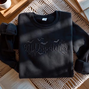 Op de afbeelding: Een zwart sweatshirt met ronde hals en geborduurde tekst 911 Dispatcher in een cursief lettertype. Het ontwerp bevat kleine geborduurde vleermuizen en spinnenwebmotieven rondom de tekst, allemaal uitgevoerd in bijpassend zwart garen.