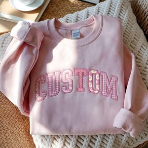 Peut inclure: Un sweat-shirt à col rond rose clair avec le mot CUSTOM en grandes lettres de style bloc sur la poitrine. Les lettres présentent un remplissage à motifs alternant des designs floraux, à pois et à carreaux vichy dans des tons de rose et blanc.