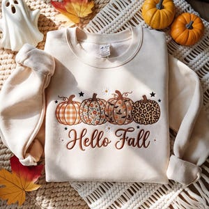 May include: A beige crewneck sweatshirt featuring a graphic of four pumpkins with different patterns, including plaid, floral, Halloween icons, and leopard print. The text "Hello Fall" is printed below in brown script, accented by small white daisies.
